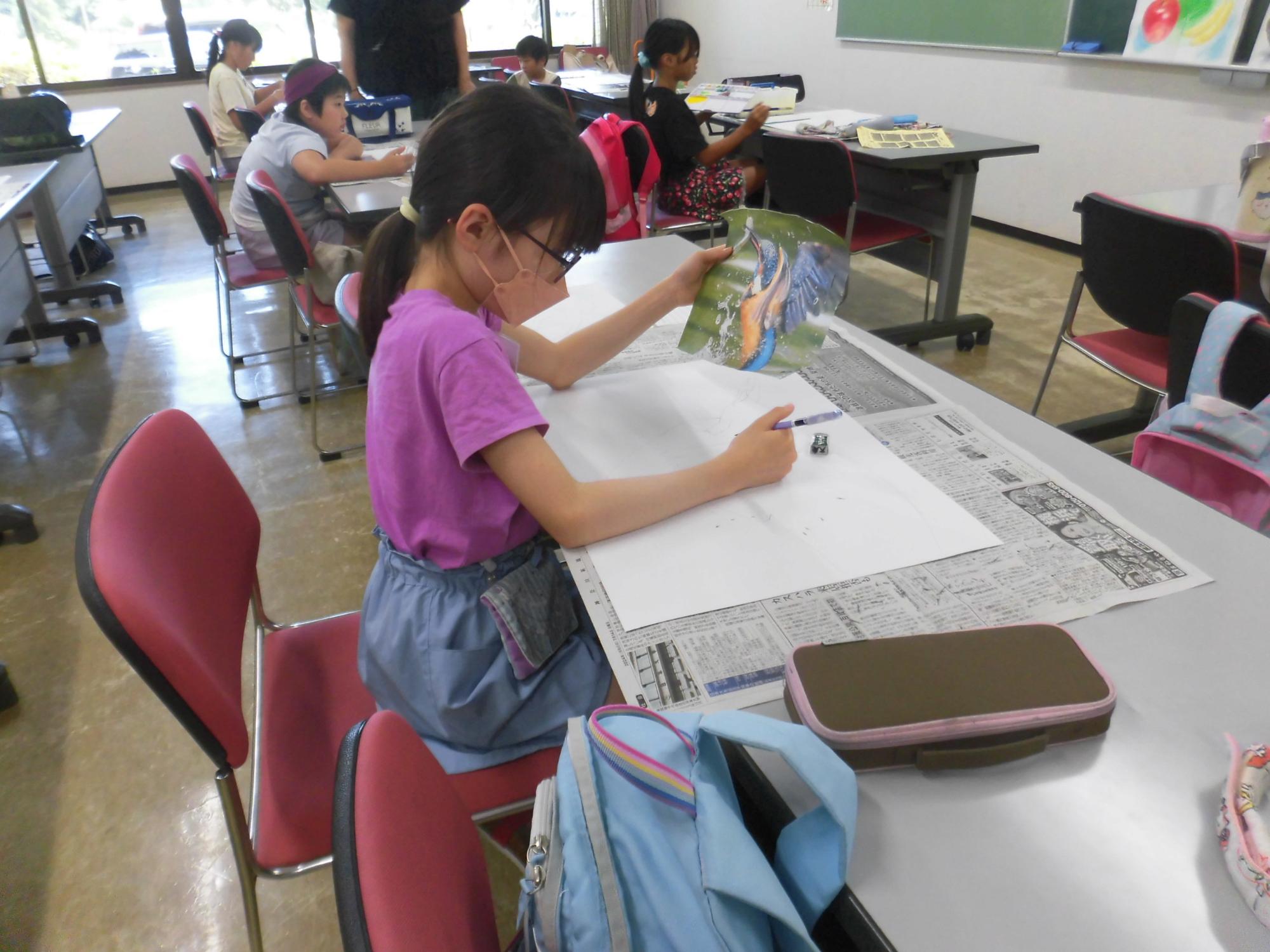 椅子に座ってメガネをかけた女の子が、大きな白い紙に鉛筆で絵を描いており、左手には見本のイラストの紙を持っている、図工や絵画教室のような場面を捉えた写真