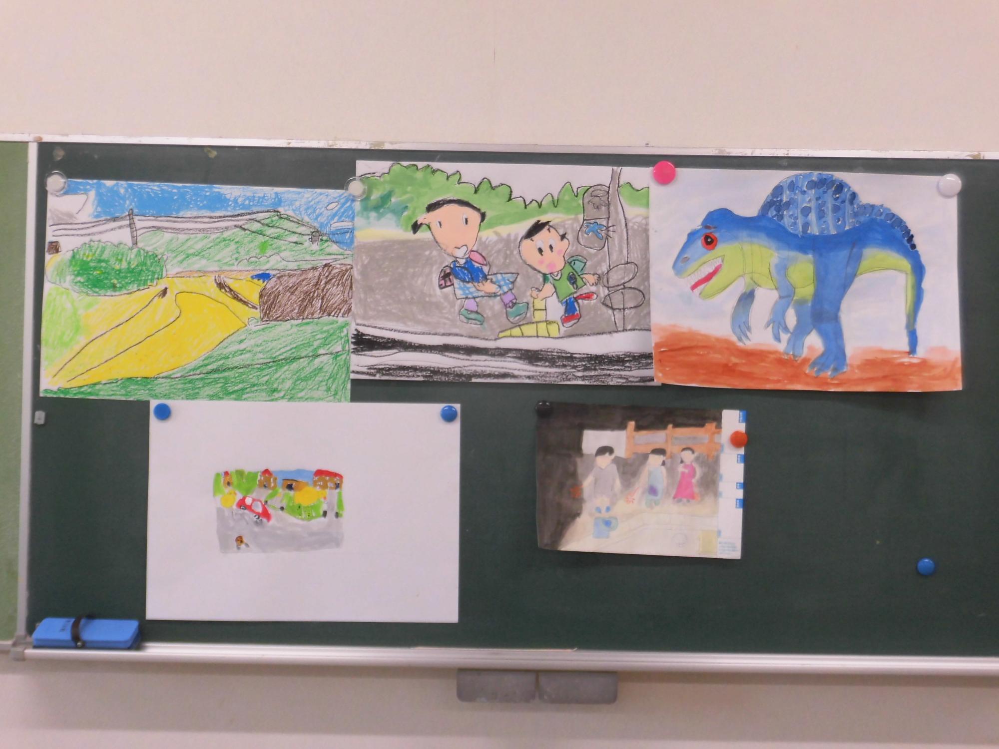 黒板に複数の画用紙が貼り付けられており、子どもが描いたカラフルな風景画や、恐竜、人物などの絵が展示されている写真