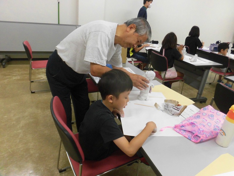 複数人の子供と大人が工作テーブルに向かい、横溝さんが子供に丁寧に作り方を教えているワークショップの写真