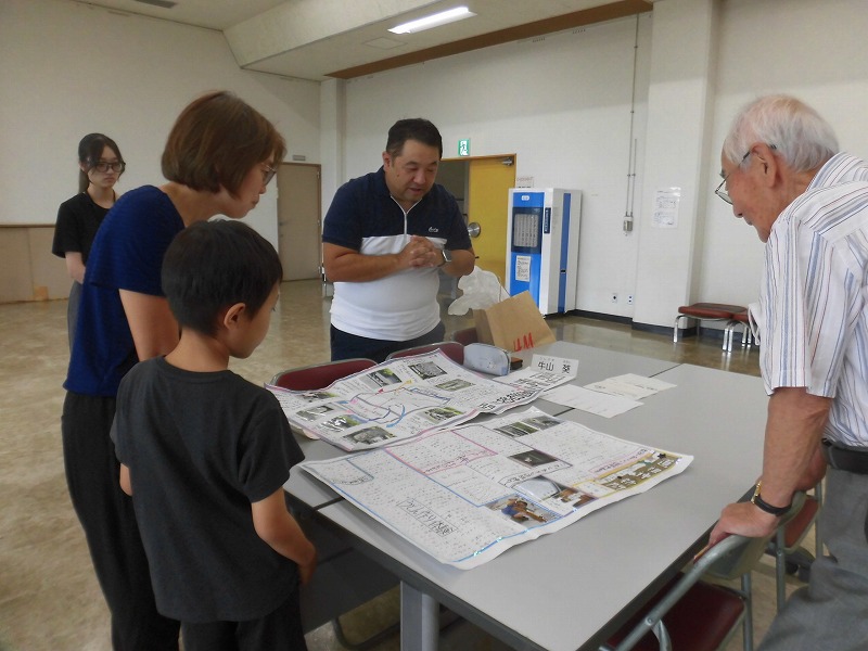 広い部屋の中で、中央のテーブルを囲んで大人4名と子供1名が、広げられた地図や写真を含む手書きの資料を覗き込み、男性が説明している様子の写真