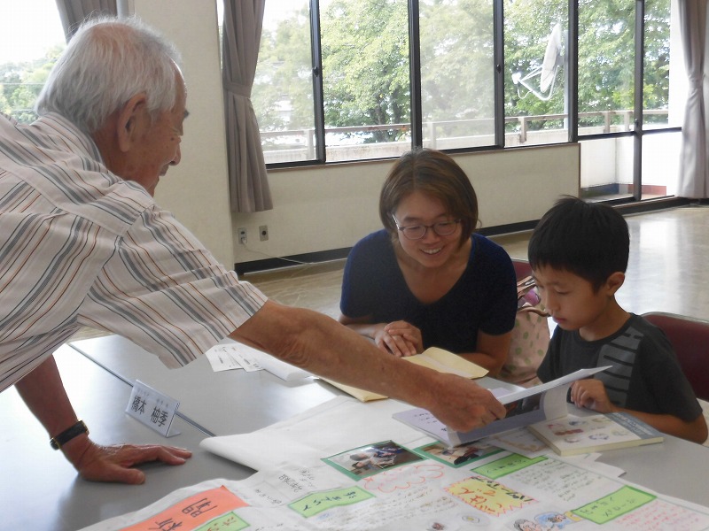 大きな部屋で武さんが、女性と男の子に資料を見せながら何かを説明している様子の写真