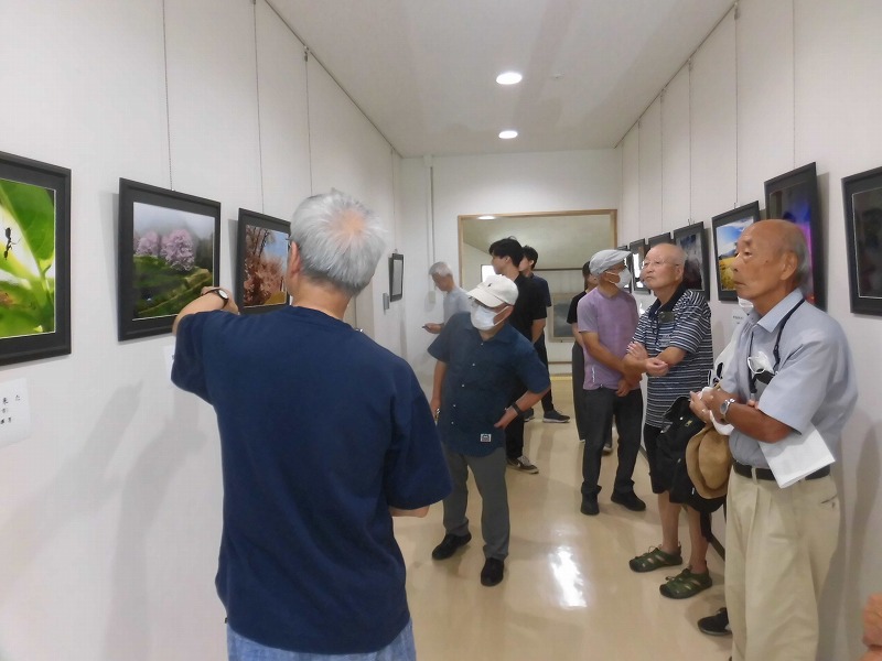 高齢の男性グループが、壁に掛けられた自然や風景などの写真を鑑賞しており、そのうちの一人が熱心に解説をしている様子の写真