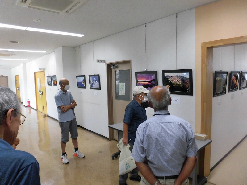 建物の廊下で数人の人々が壁に飾られた風景写真などを鑑賞しており、そのうちの一人が作品を指差しながら熱心に話している、屋内で行われた写真展の様子の写真