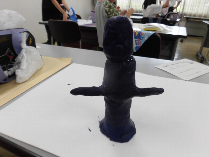 暗い青色の粘土で制作された、両腕を水平に広げた人型の抽象的な立体作品が机の上に置かれている写真