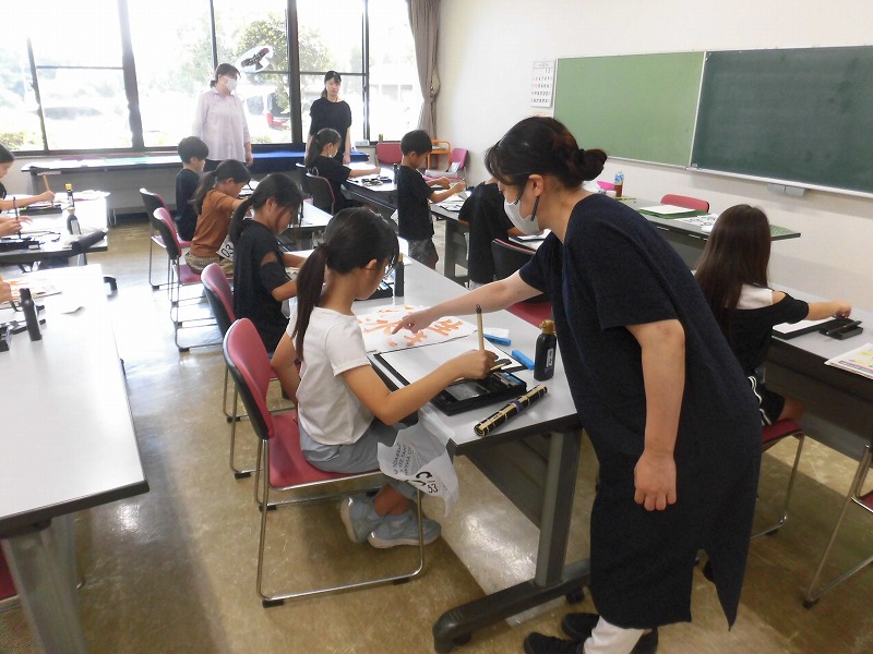 複数の子供たちが机に向かって書道に取り組んでおり、マスクを着用した田中寛絵さんが一人ひとりに丁寧に指導している、書道教室の写真