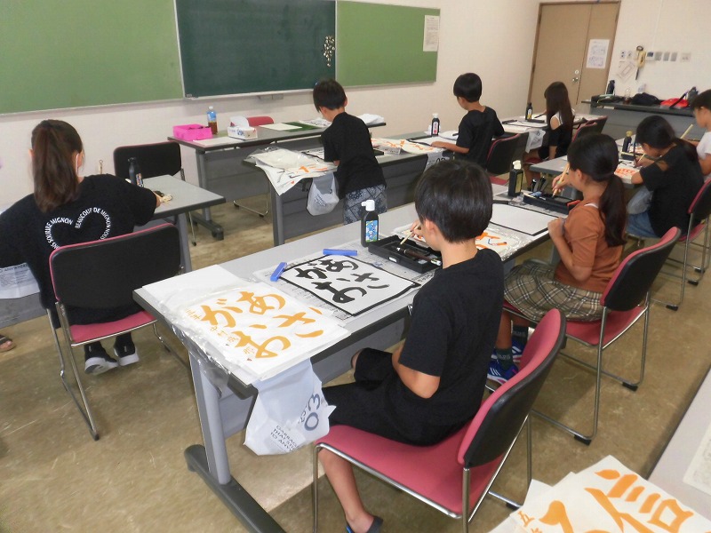椅子に座った子供たちが筆や墨を使い、机の上に広げた半紙に文字を書いている、学校の教室で行われている書道教室の写真