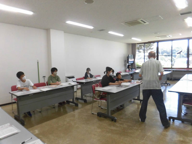 テーブル席に座った子供と大人たちが、部屋の前に立って説明する田中淑生さんに注目している様子の写真