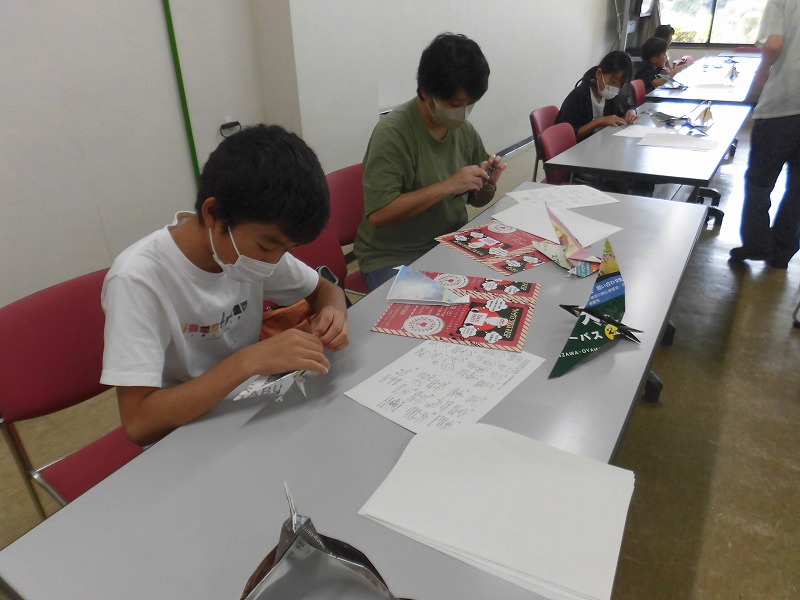 数人の子供と大人がテーブルに向かい、折り紙や紙の工作を熱心に行っている様子の写真