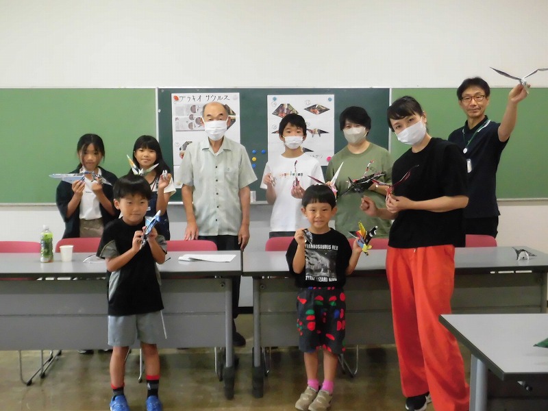 家族や子供たちが教室で自作の紙飛行機を持ち、黒板を背景に笑顔でカメラに向かって集合している写真