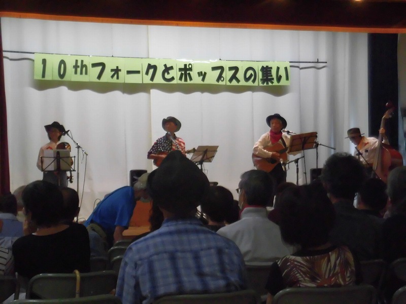 ステージの上で4人組のバンドが楽器を演奏し、観客が客席からその様子を鑑賞しており、「10thフォークとポップスの集い」という横断幕が掲げられている、屋内の音楽イベントの写真