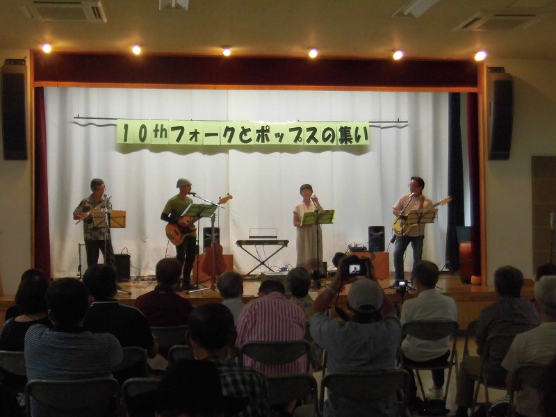 「10thフォークとポップスの集い」という横断幕が掲げられたステージで、4人組のバンドが楽器を演奏し、観客がその様子を携帯電話やカメラで撮影しながら鑑賞している様子の写真