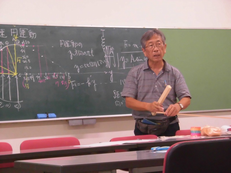 教室で木村晴生先生が黒板の前に立ち、物理学の数式や図が書かれた黒板を背景に、木製の道具を持って講義をしている様子の写真