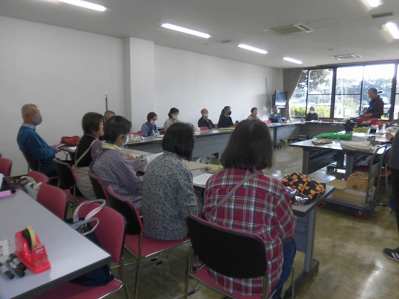 テーブルに向かい合って座る多くの参加者が、一人の講師から説明を聞いている様子の写真