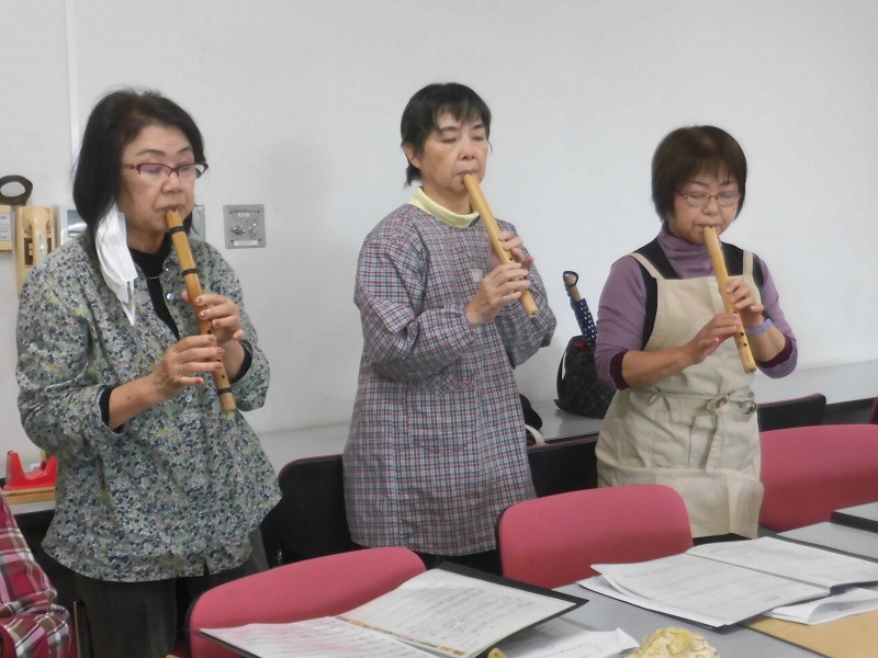 部屋の中で3人の女性がそれぞれのマイケーナを持ち、楽譜を見ながら一斉に演奏している様子の写真