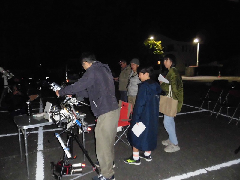 夜空の下、数人の人々が天体望遠鏡や機材が並んだ場所で、天体観測会に参加している様子の写真