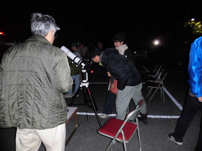 夜間に屋外で、複数の参加者が集まり、一人が望遠鏡を覗き込んでいる、天文観測イベントの様子の写真
