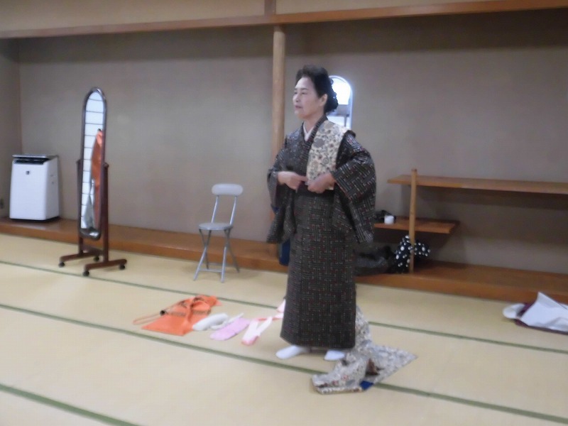 女性が鏡の前に立ち、着物を着付けしており、床には扇子や着付け小物が置かれている、着付け教室か着物体験会の写真