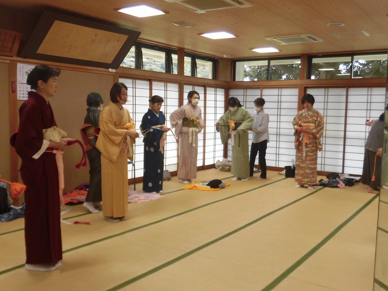 畳敷きの広い和室で、複数人の女性たちが着物を着付けしており、互いに手伝いながら和装の練習をしている様子の写真