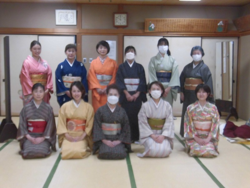 華やかな着物を着た複数の女性たちが和室に集まり、笑顔でカメラの方を向いて記念撮影をしている様子の写真