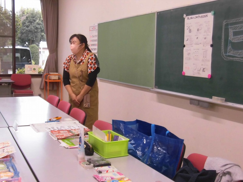 教室内でエプロンを着用した川口京子さんが、黒板に貼られたトートバッグの作り方の説明書きを背景に、机に並べられた道具類とともに参加者を待っている様子の写真
