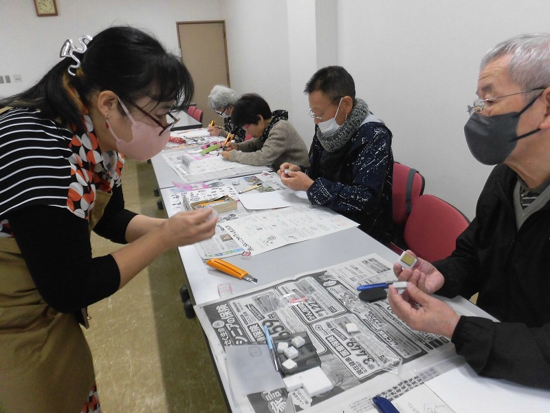 教室内でマスクを着用した男女が、机に新聞紙を敷き、カッターナイフや道具を使いながら、川口京子さんから彫刻の指導を受けている様子を捉えた写真