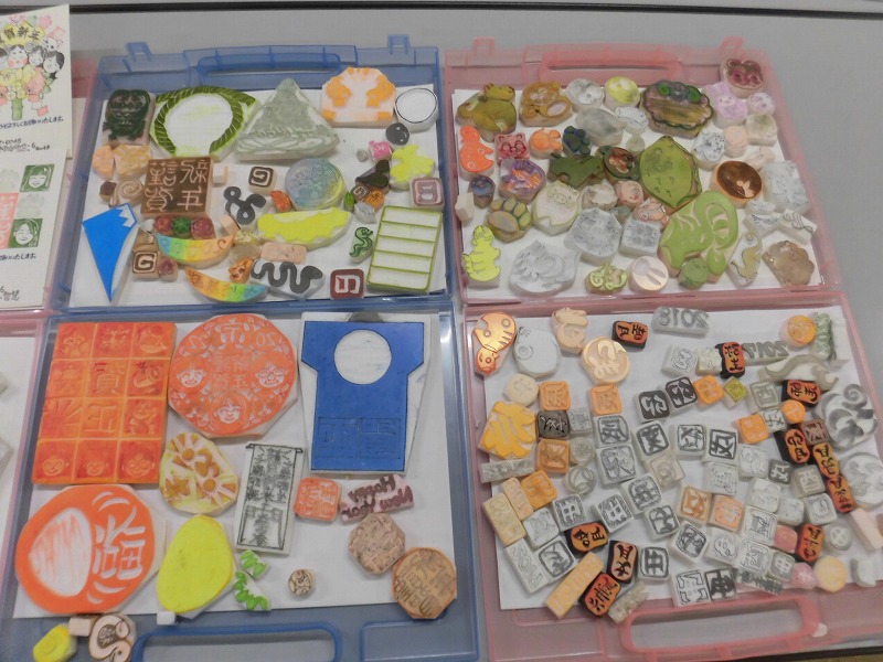4つのプラスチックケースに整理された多数の消しゴムはんこが並んでおり、動物や文字、日本の縁起物など様々なモチーフの手作り作品が収められている写真
