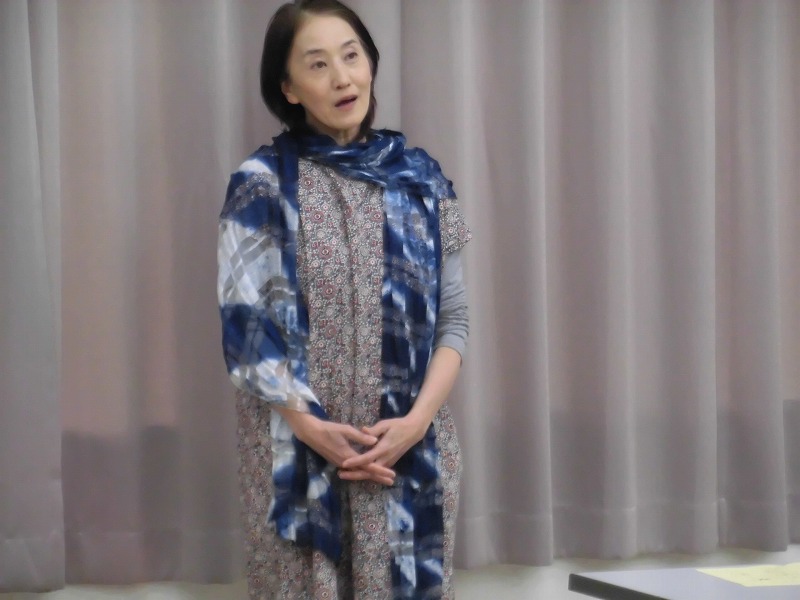 水色のスカーフを首に巻いた三溝美香さんが、何かを語りかけているように口を開け、聴衆の前に立っている姿を捉えた発表会の写真