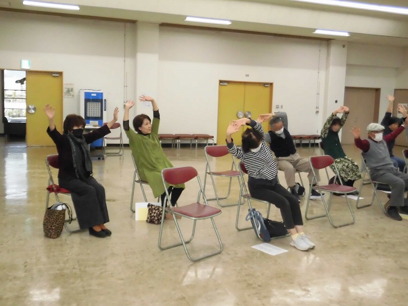 椅子に座った複数の参加者が両手を上げて体を横に傾けるストレッチ体操をしている、健康増進教室または体操教室の写真