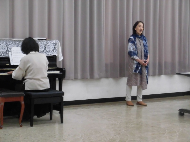 広々とした室内で、一人の女性がピアノを弾き、もう一人の三溝美香さんが立って歌っている、発表会か練習風景を写した写