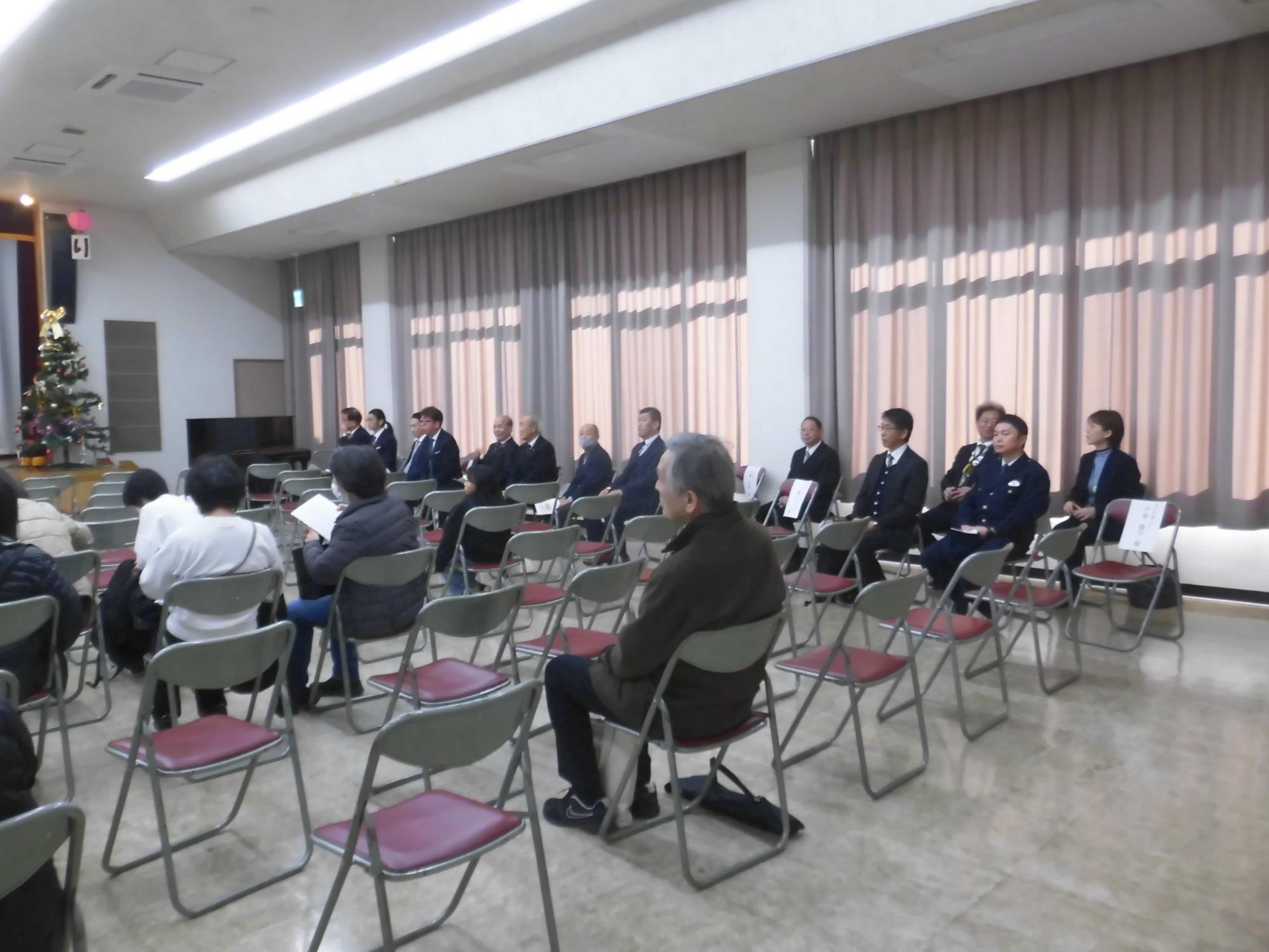 白い壁とグレーのカーテンがある明るいホールで、スーツ姿の多数の参加者が赤い座席の折りたたみ椅子に座り、前方に立つグループに向かって式典に参加している様子の写真