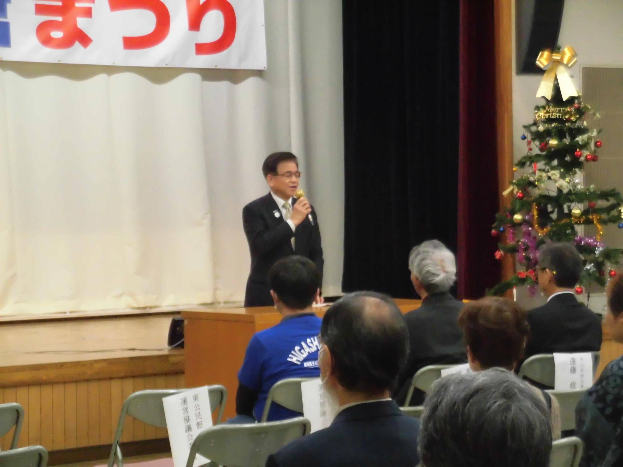 白いカーテンと赤い横断幕、右側にクリスマスツリーが飾られたステージで、スーツ姿の市長がマイクを持って話し、多数の聴衆が座っている聞いているイベント会場の写真