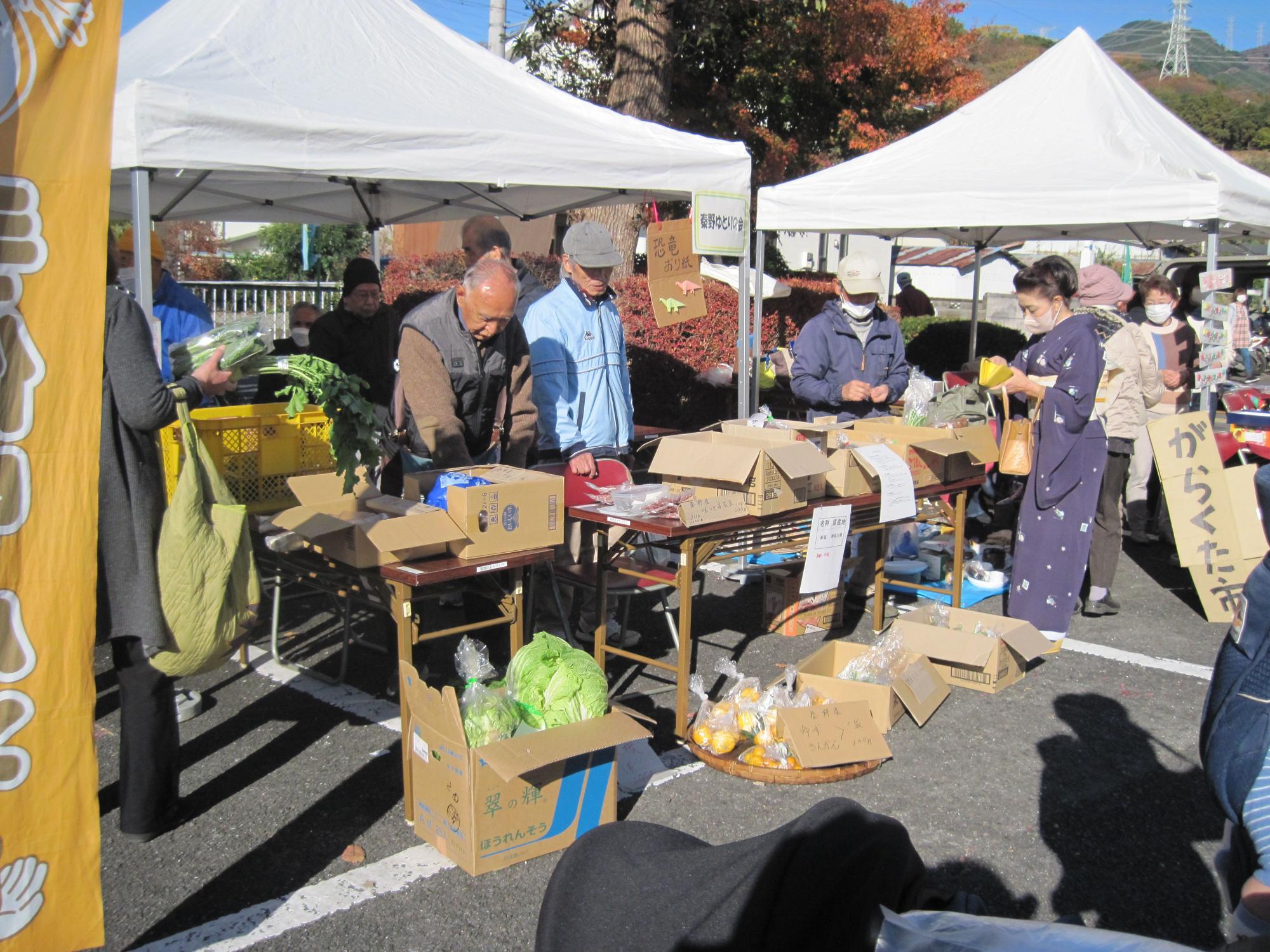 青空の下、白いテントが複数張られた屋外イベント会場で、木製テーブルに野菜や商品が並べられた段ボール箱が置かれ、多数の参加者が農産物や食品を手に取りながら買い物をしている様子の写真