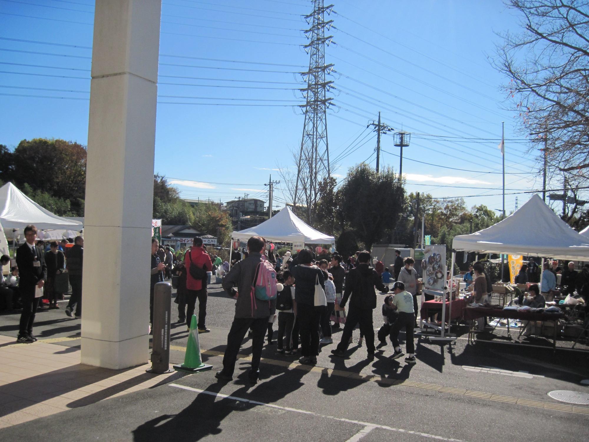 青空の下、白い支柱のある舗装された広場で、複数の白いテントが設置され、多数の参加者が歩き回ったりテーブルの周りに集まったりしている屋外イベントの写真