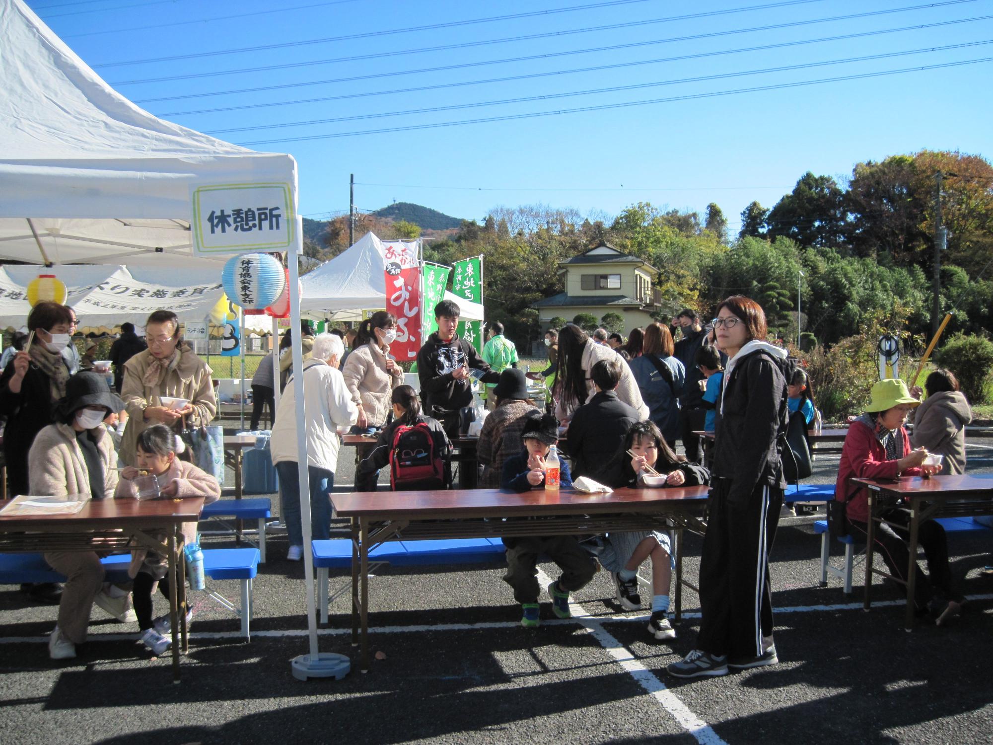 青空と緑の木々を背景に、白いテントと「休憩所」の看板が設置された屋外イベント会場で、多数の参加者が木製テーブルとベンチに座って食事をしたり歩き回ったりしている東公民館祭りの写真