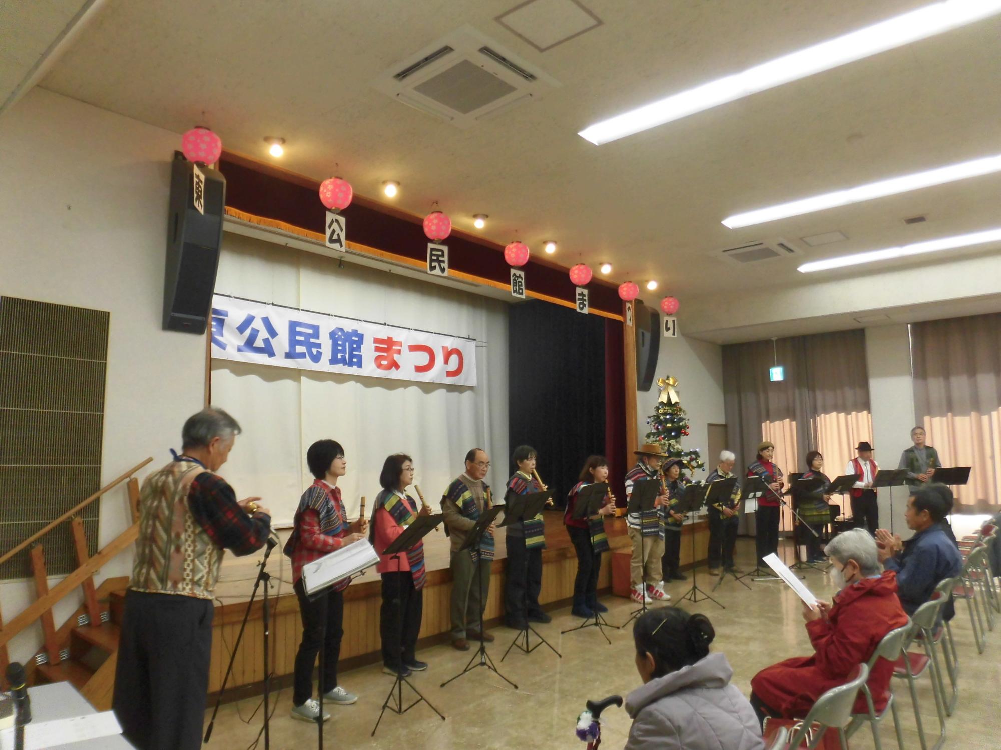 白いカーテンに「公民館まつり」と書かれた横断幕が掲げられたステージで、カラフルな衣装を着た10名以上の演奏者が楽器を手に並び、前の客席には数名の観客が座って鑑賞している写真