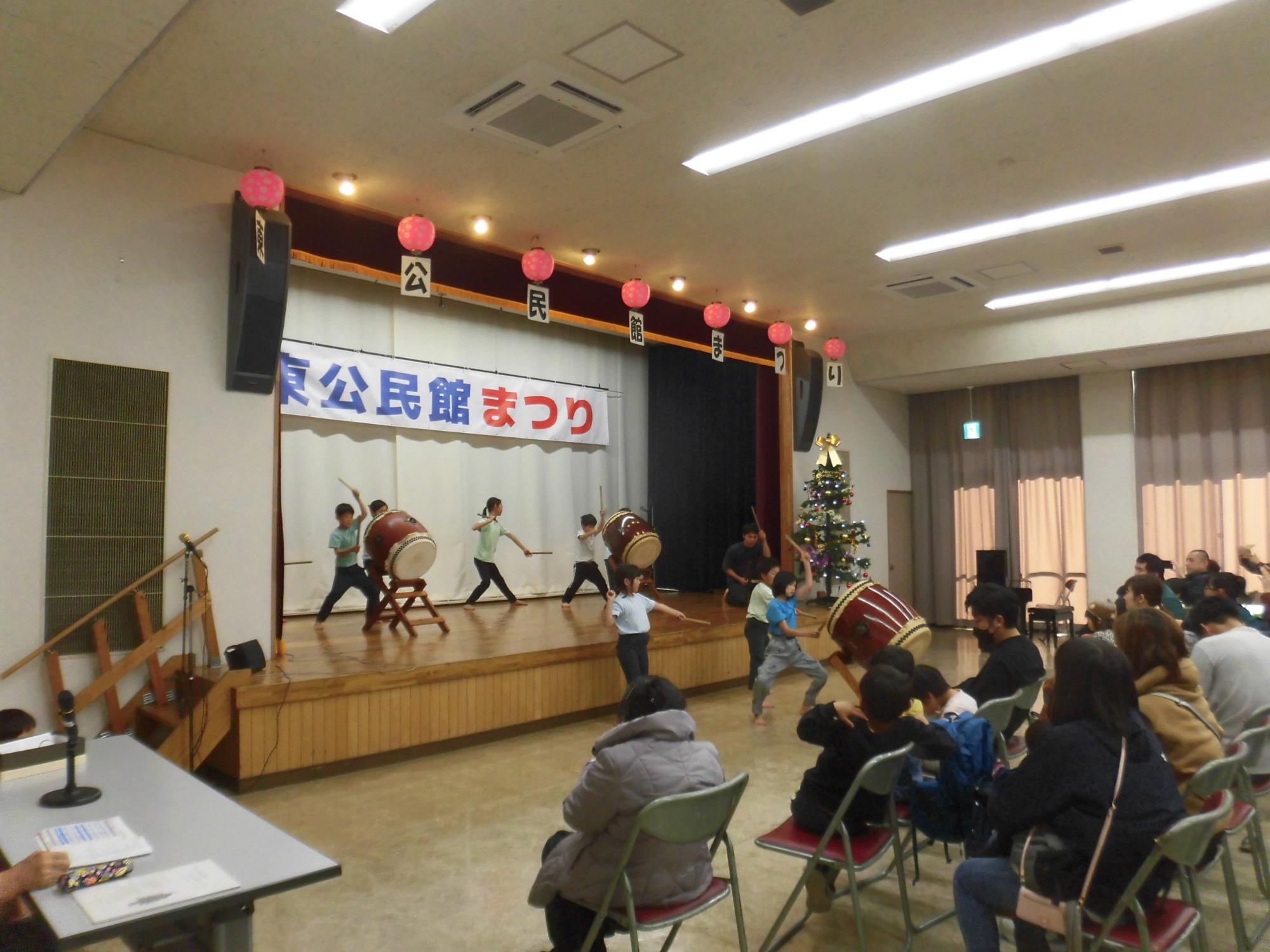 白いカーテンに「公民館まつり」と書かれた横断幕が掲げられたステージで、白と青の衣装を着た複数の演奏者が大きな太鼓を叩き、下の客席には多数の観客が座って鑑賞している写真