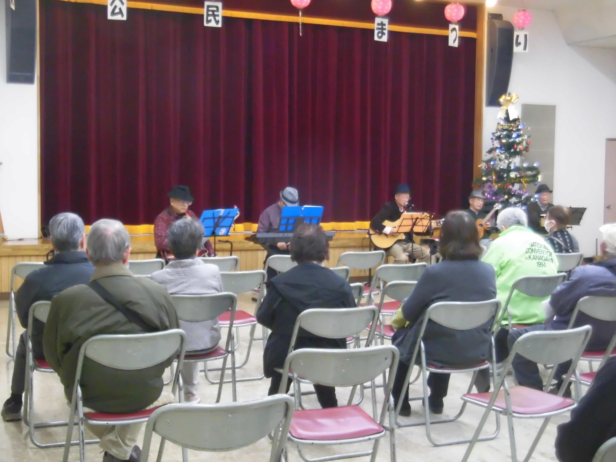 赤紫色のカーテンがあるステージで、帽子をかぶった5名の演奏者が青い譜面台を前に楽器を演奏し、下の客席には多数の観客が座って鑑賞している写真