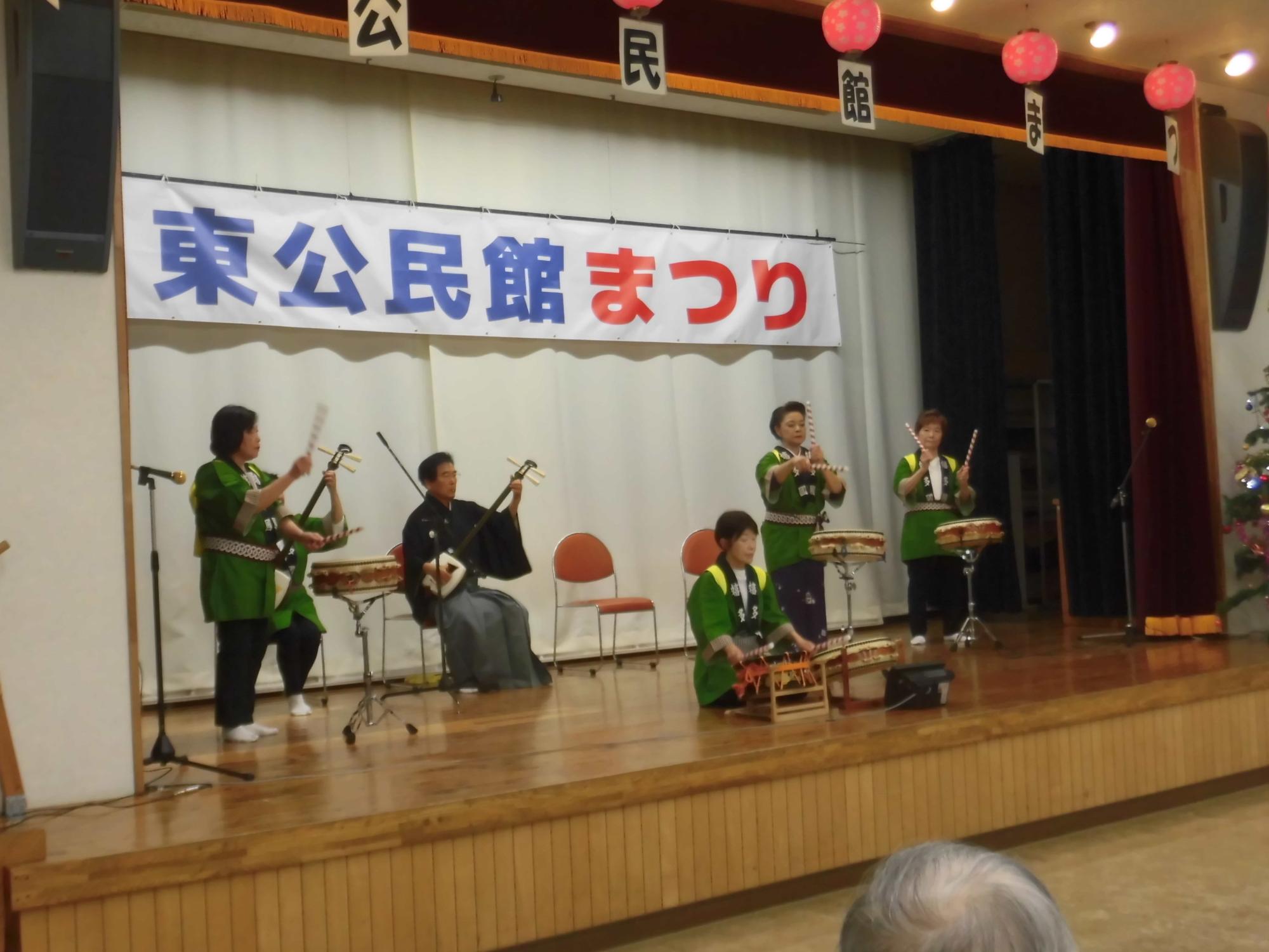 白いカーテンに「東公民館まつり」と書かれた横断幕が掲げられた木目調のステージで、緑と黒の衣装を着た5名の演奏者が三味線などの和楽器を演奏している写真