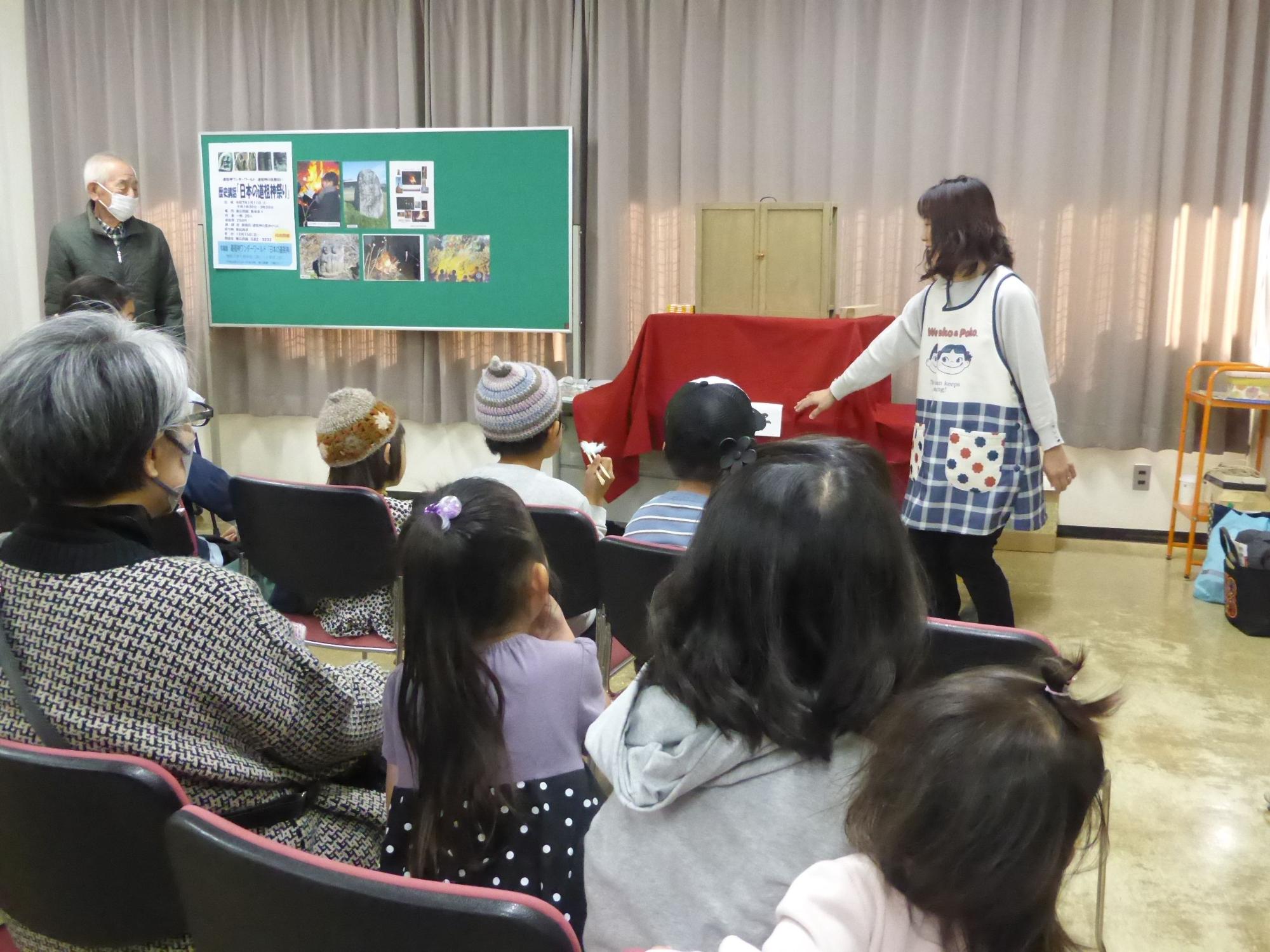 白いカーテンのある室内で、エプロンを着た女性が赤い布の上に置かれた紙芝居を使って、椅子に座った子どもたちと高齢者に読み聞かせをしている写真