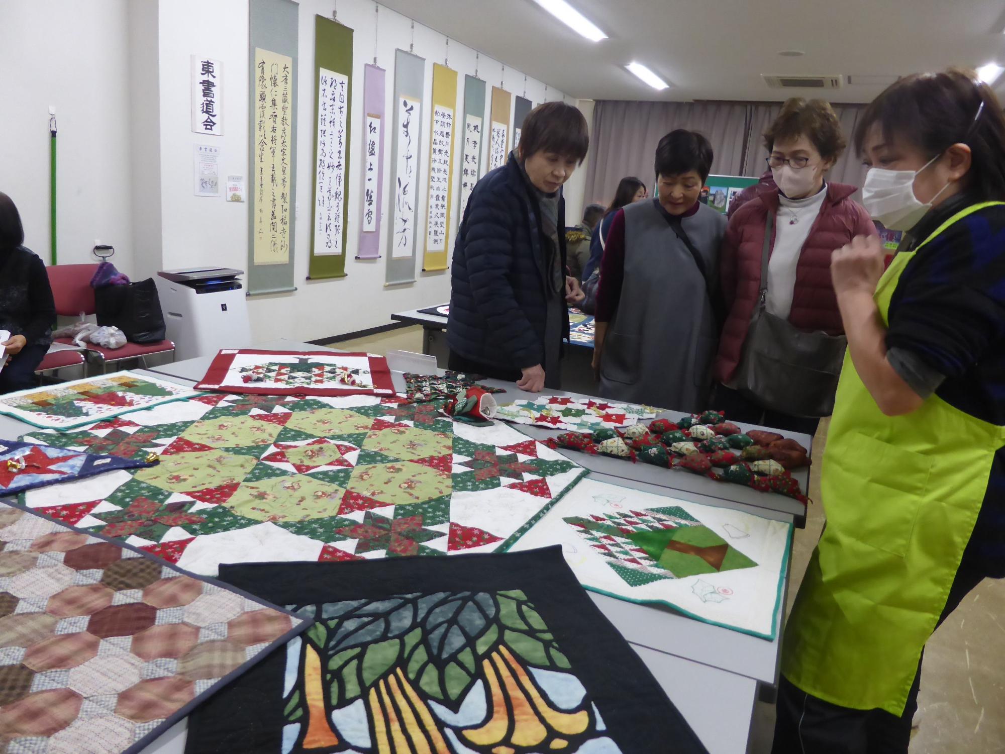 白い壁に複数の書道作品が掲示され、長机の上にクリスマス柄のパッチワークキルトや手芸作品が展示された室内で、複数の参加者が作品を鑑賞している写真