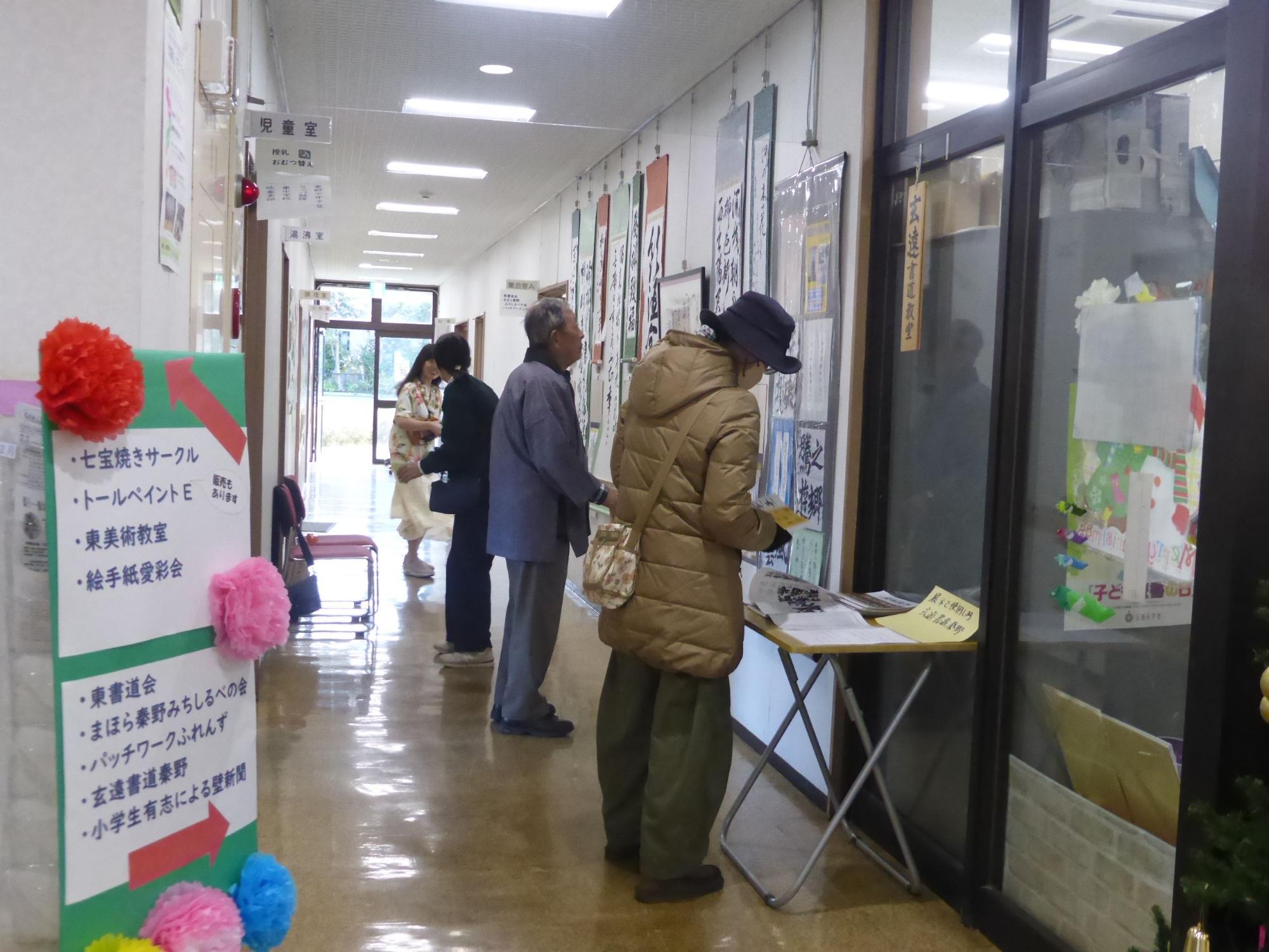 白い壁に多数の書道作品が掲示された明るい廊下で、複数の参加者が展示作品を鑑賞している様子の写真