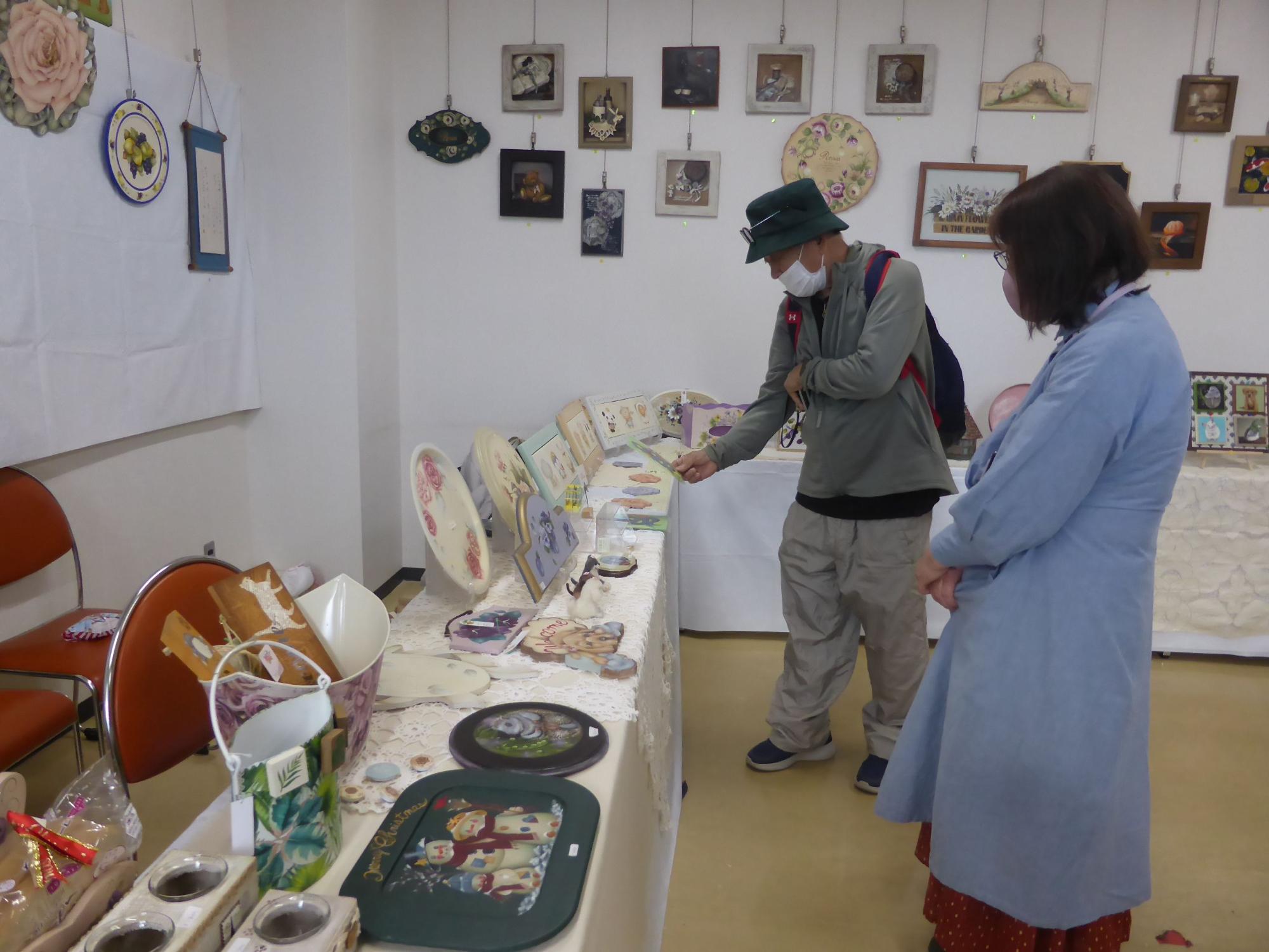 白い壁に多数の絵画や工芸品が掲示され、手前の白い展示台にはトールペイント作品やバッグ、皿などの手芸品が並べられた展示会場で、2名の参加者が作品を鑑賞している写真