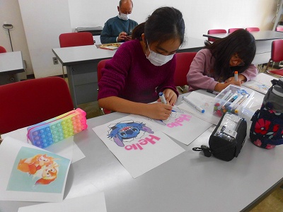 白い部屋の机で、マスクをつけた二人の少女が、ポップなキャラクターの絵にマーカーペンで色を塗るワークショップに参加している様子の写真