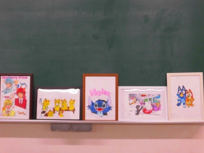 黒板の下の棚に並べられた子どもたちのイラスト作品の展示で、それぞれの額縁には「ステッチ」や「ブルーイ」、「ストロベリープリンス」などの人気キャラクターが描かれている様子の写真