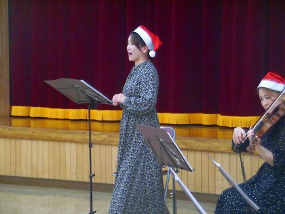 赤い幕が背景のステージで、サンタの帽子をかぶった女性二人が、楽譜を見ながら一人は歌を歌い、もう一人はバイオリンを弾いているクリスマスコンサートの様子の写真