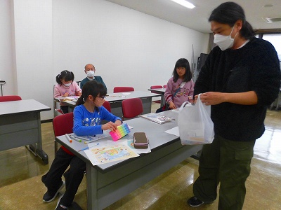 机を並べた室内で、複数の子どもたちが色塗りワークショップに参加しており、一人の男性が子どもたちに紙袋を手渡している様子の写真