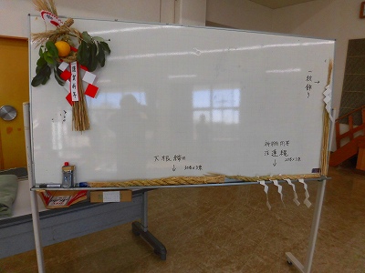 左上には橙と稲穂が飾られ、板面には「神棚用」「注連縄」と書かれ、下に藁が置かれた、しめ縄作りの講習会で使われたホワイトボードの写真