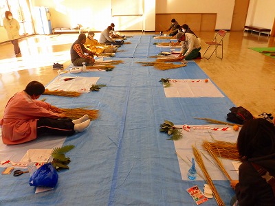 青いシートが敷かれた広間で、マスクを着用した数人の参加者が、稲わらや葉、橙を使って、新年を迎えるためのしめ縄作りに熱中している様子の写真