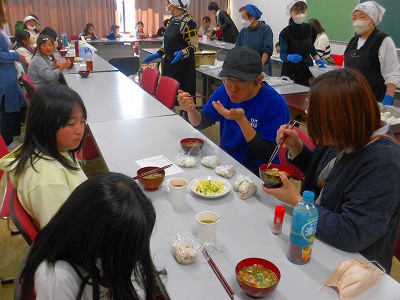 広い部屋の机を囲んで、複数の参加者が料理を試食しており、それぞれの前にはおにぎりや味噌汁などが並べられ、和気あいあいとした雰囲気の中で食事を楽しんでいる様子の写真