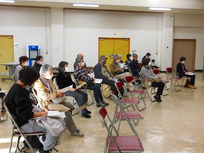 広い室内で、マスクを着用した聴講者が、茶色い折りたたみ椅子に間隔を空けて座り、手に持った資料を見ながら演奏を熱心に聴いている様子の写真